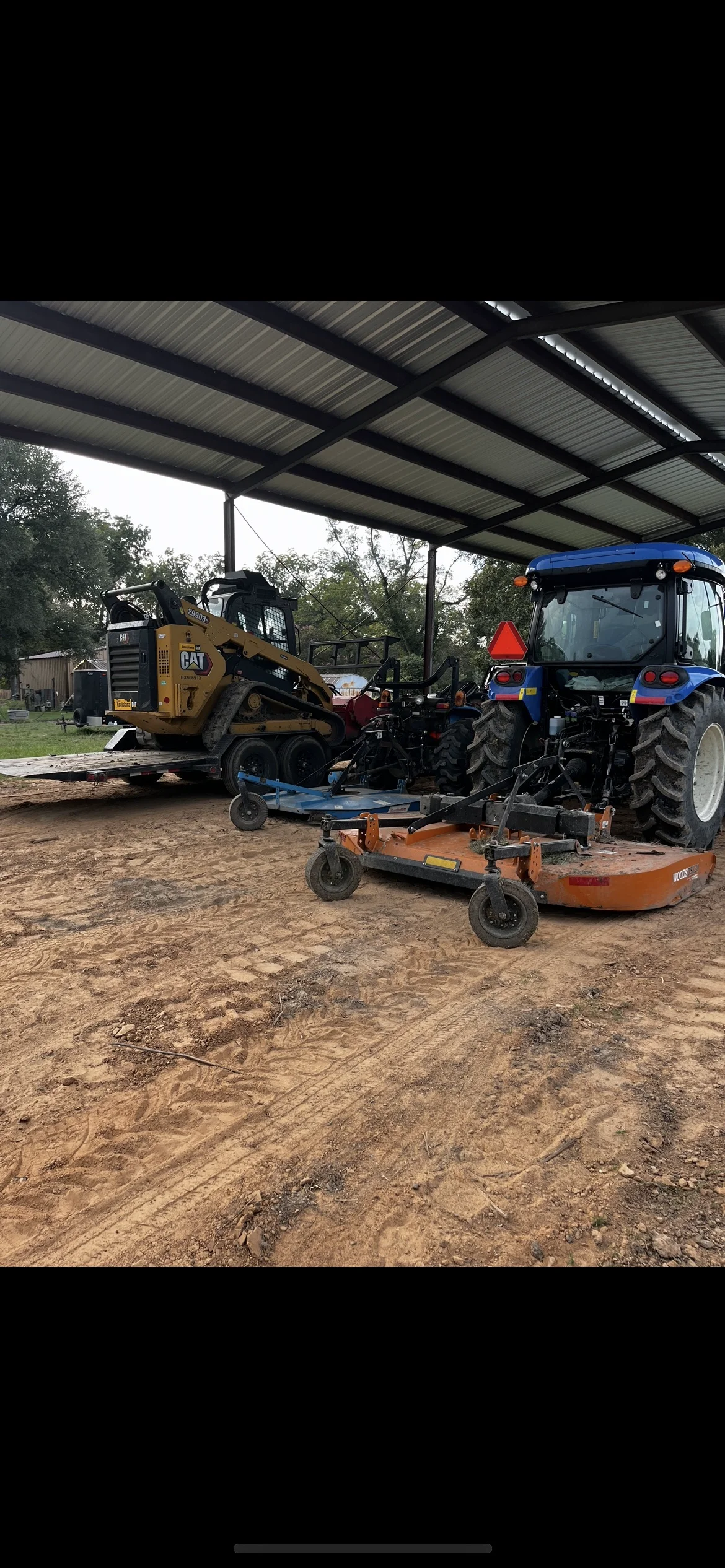 Metal equipment shelter with corrugated steel roof over compacted dirt pad, housing skid steer and tractor