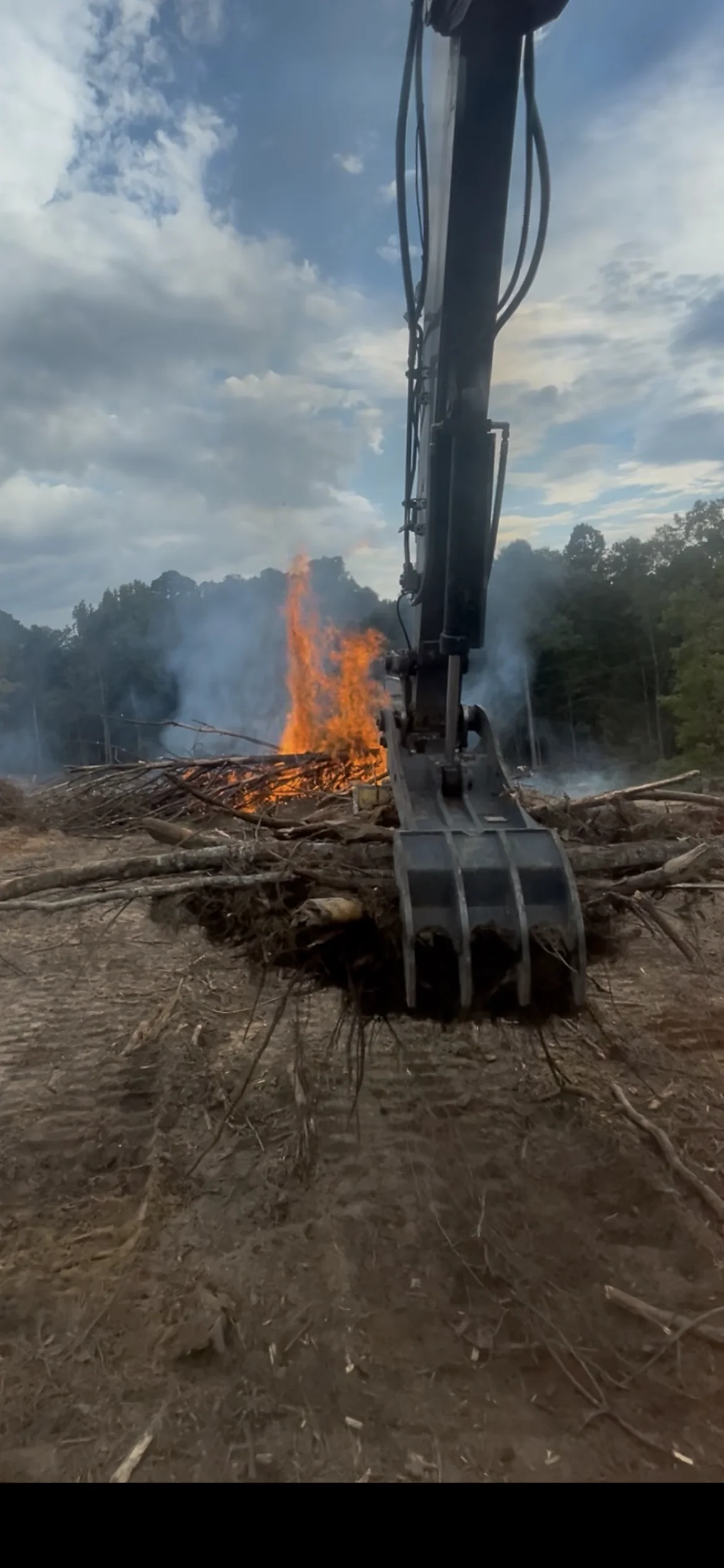Excavator bucket clearing felled logs with active debris fire burning in background