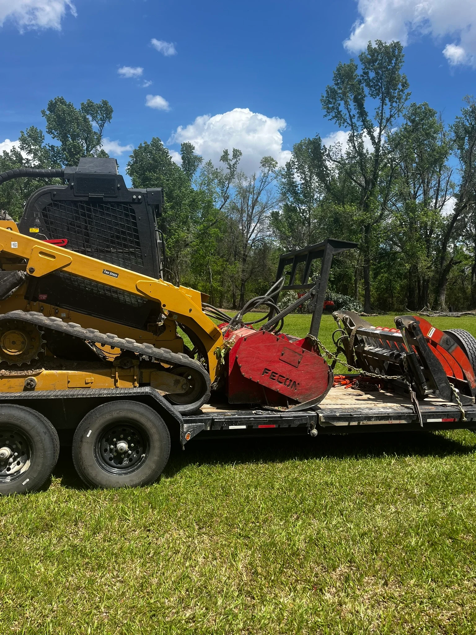 Yellow skid steer with grapple attachment loading felled trees onto trailer with red wood chipper