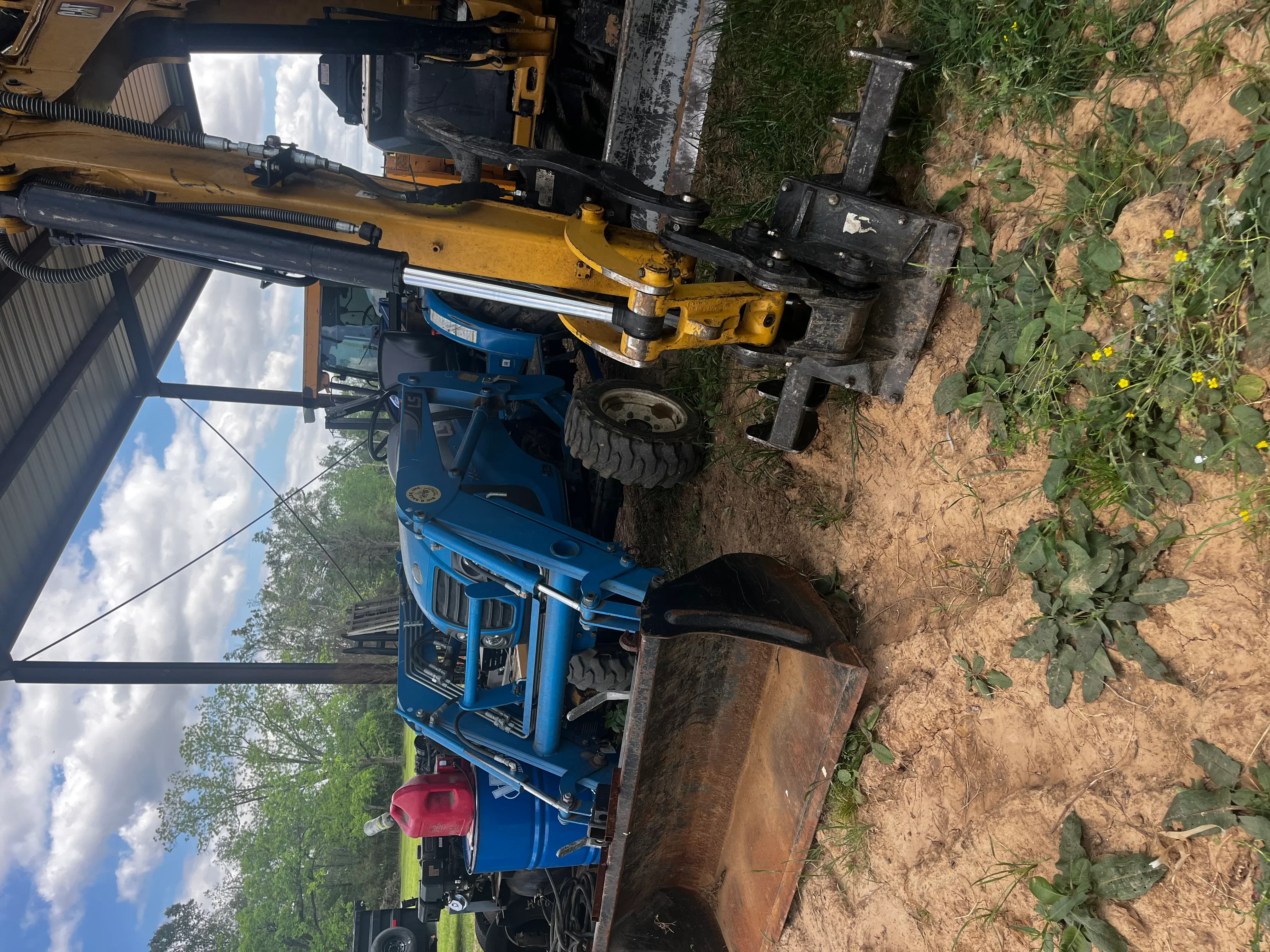 Blue excavator with yellow boom attachment clearing overgrown land with dirt and vegetation